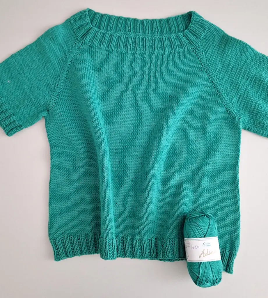 Grünes, kurzärmeliges T-Shirt aus Strick mit einem Knäuel Garn daneben.