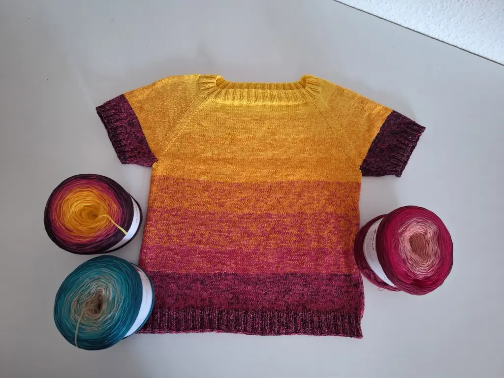 Gestreiftes Kinderoberteil in Gelb, Lila und Pink, umgeben von drei Garnknäueln.