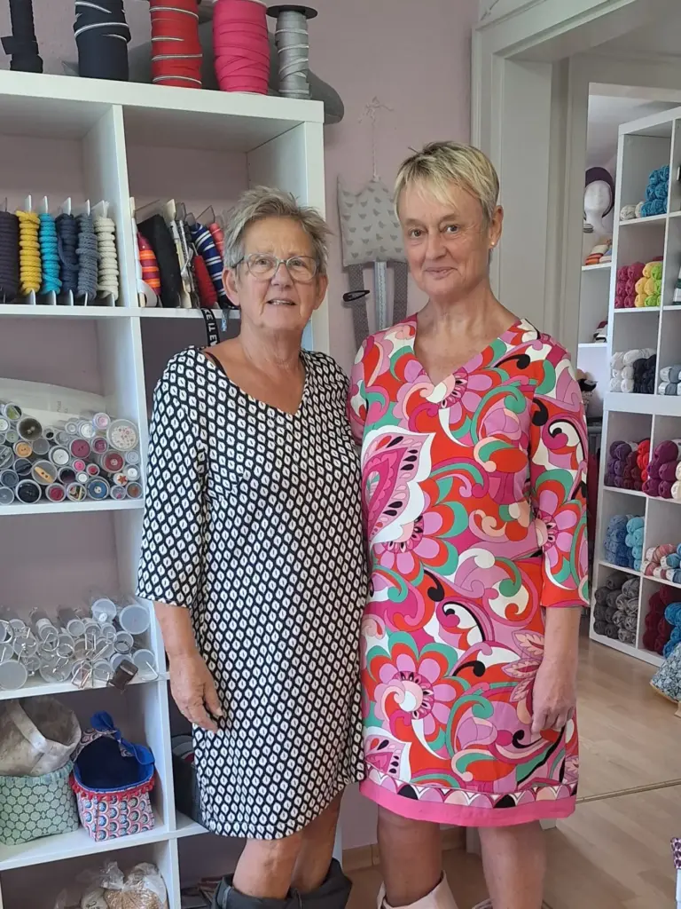 Zwei Frauen stehen nebeneinander, eine trägt ein schwarz-weißes Kleid, die andere ein buntes Kleid mit floralem Muster.