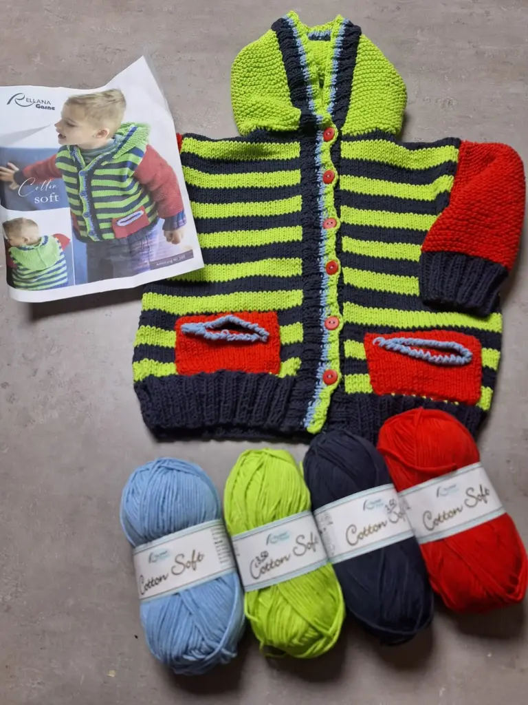 Gestreifte Kinderjacke in Grün, Blau und Rot mit Kapuze, daneben ein Strickmusterheft und vier Wollknäuel in Blau, Grün, Dunkelblau und Rot.