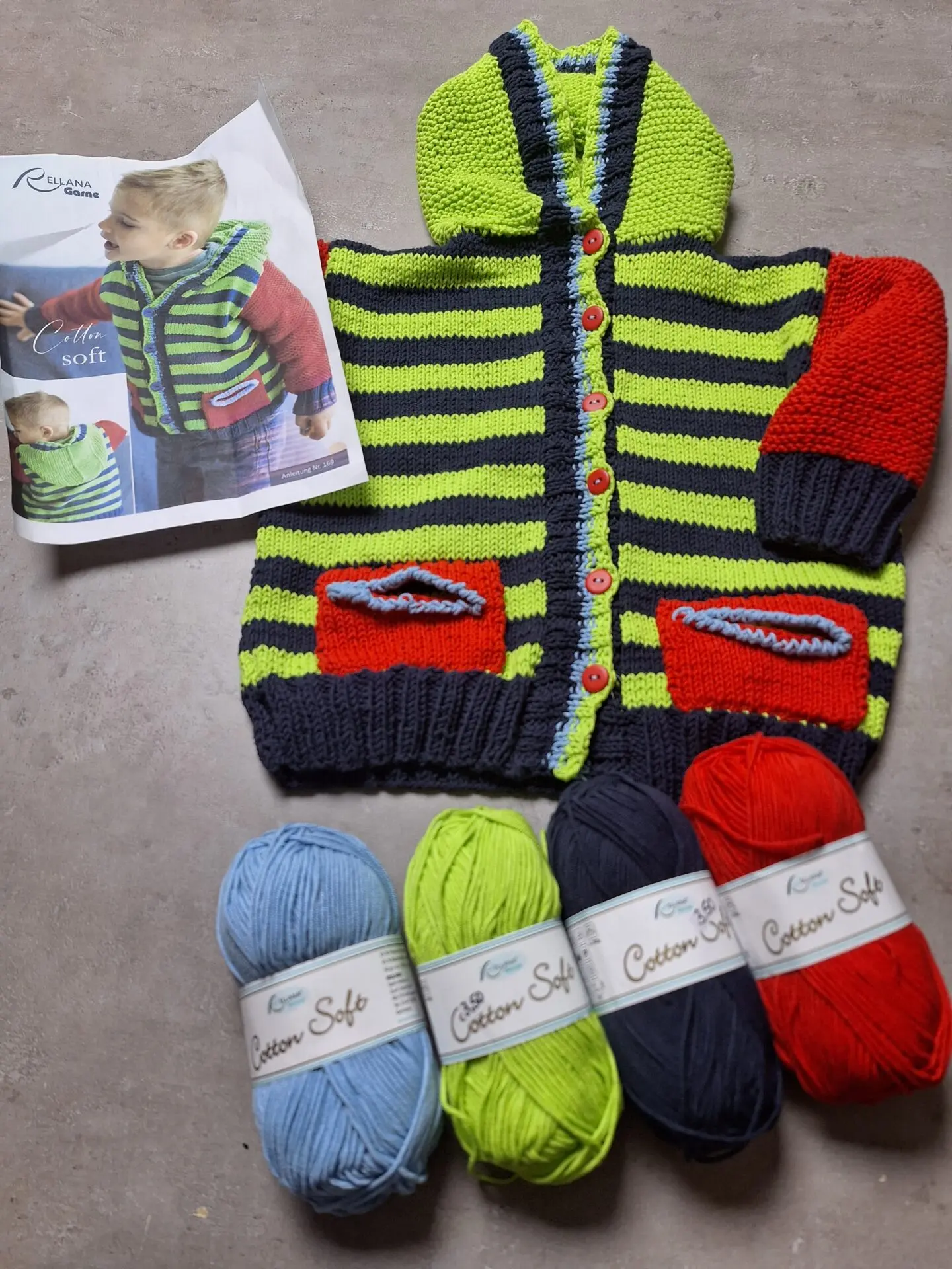 Gestreifte Kinderjacke in Grün, Blau und Rot mit Kapuze, daneben ein Strickmusterheft und vier Wollknäuel in Blau, Grün, Dunkelblau und Rot.