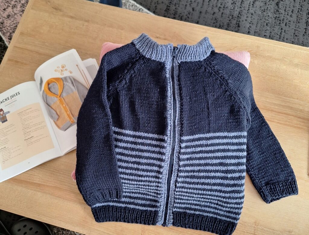 Kinderjacke aus Wolle in Blau mit grauen Streifen und Rundhalsausschnitt, liegend auf einem Tisch neben einer Strickanleitung.