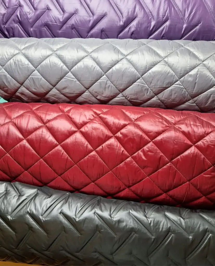 Gestapelte, gesteppten Stoffrollen in lila, grau, rot und schwarz mit Rautenmuster.