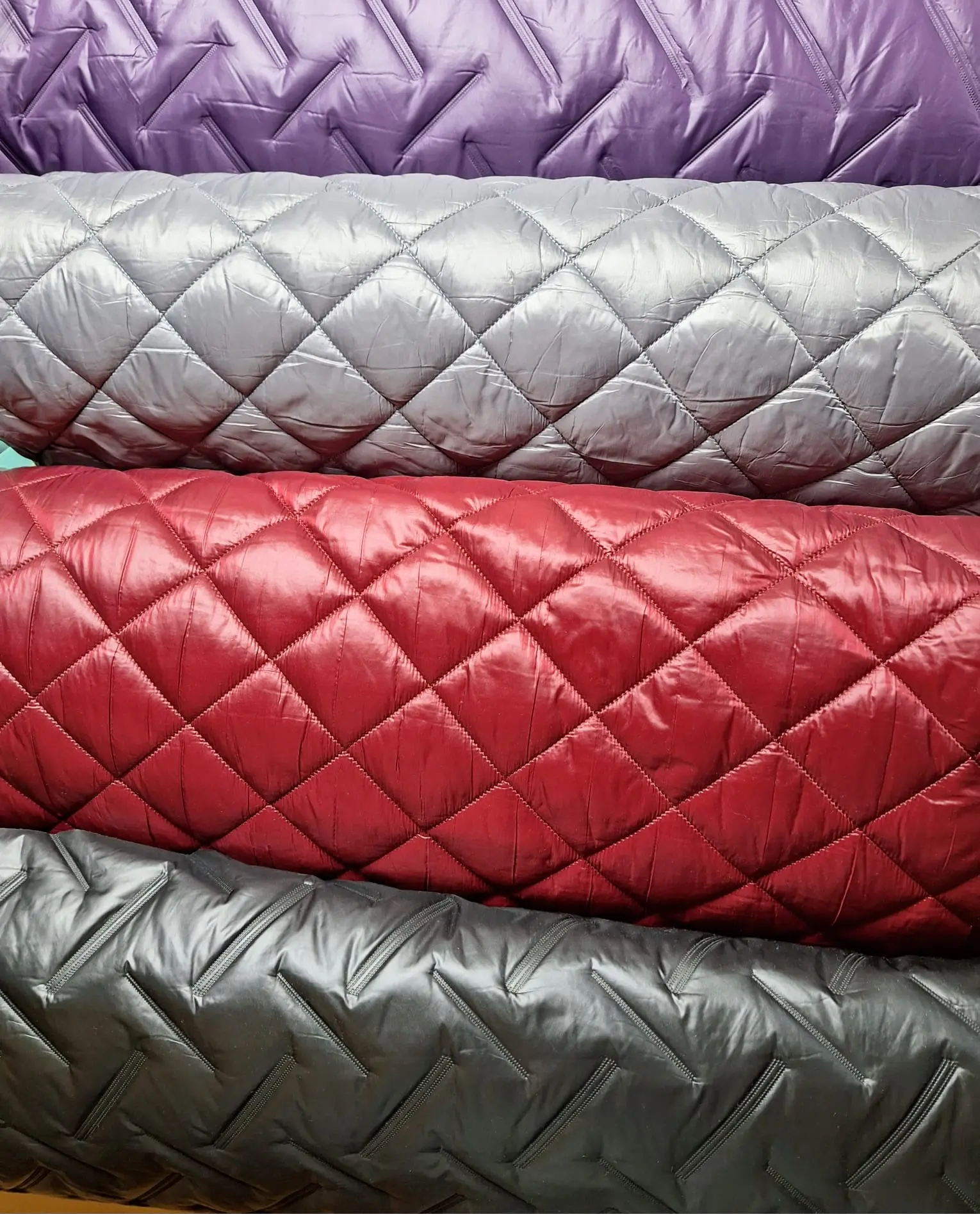 Gestapelte, gesteppten Stoffrollen in lila, grau, rot und schwarz mit Rautenmuster.