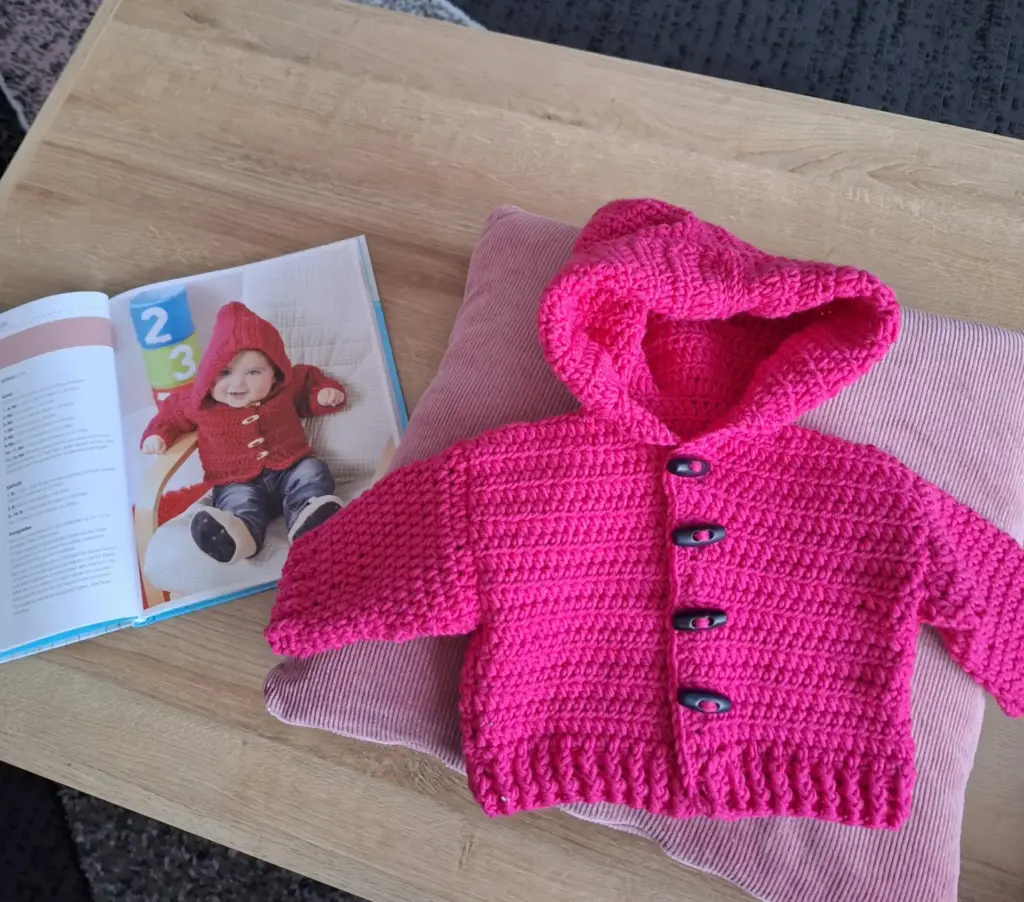 Rosa gehäkelte Jacke mit Kapuze und Knöpfen auf einem Kissen, neben einem Buch mit einem Modell in ähnlicher Kleidung.